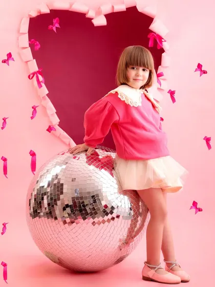 A stylishly dressed girl poses with a large disco ball in a pink, heart-themed Valentine's setup. This image combines retro fun with modern style for a unique kids' portrait.