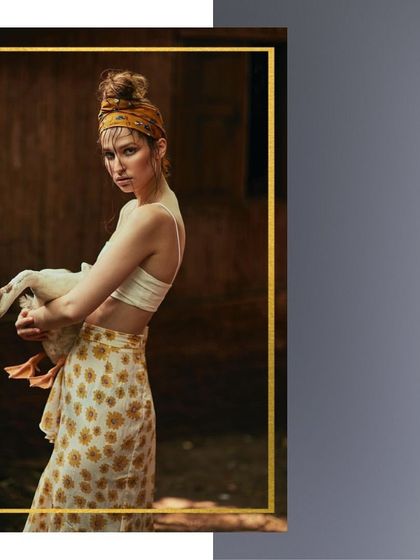 An editorial shot that blends rustic simplicity with high fashion. The styling is bohemian and relaxed, with a printed skirt and headband, while the unusual prop of a duck adds a quirky, narrative element.