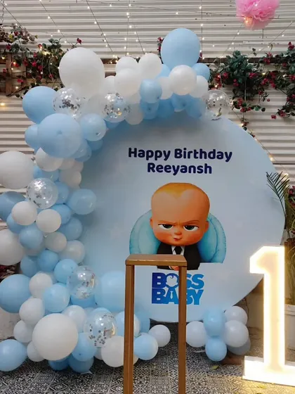 A Boss Baby first birthday decoration for Reeyansh. This outdoor-friendly setup features a custom backdrop, a blue and white balloon garland with confetti balloons, and a light-up number '1'.