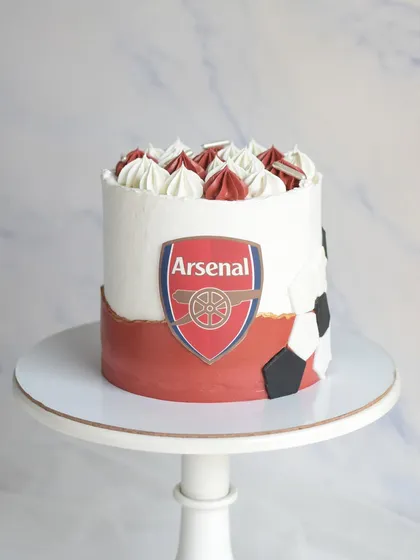 For an Arsenal football club fan, this cake features the team's logo and colors. The design includes a soccer ball pattern on the side and is topped with red and white frosting swirls.