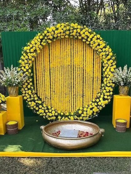 A classic Haldi backdrop for outdoor functions. This setup features a large circular frame filled with strings of yellow marigolds, a green panel background, and a traditional brass urli in the center for the ceremony.