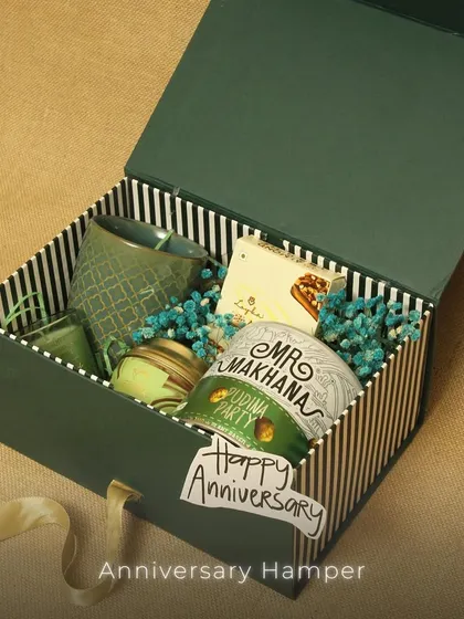 An anniversary hamper designed for a cozy celebration. This gift set is packed in a green and white striped box and includes a stylish mug, snacks like Mr. Makhana, and a scented candle.