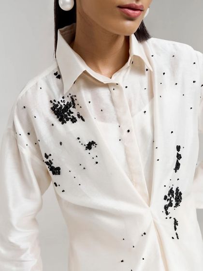 A close-up of the draped ivory shirt, showcasing the delicate black beaded embellishments that create the artistic splatter effect.