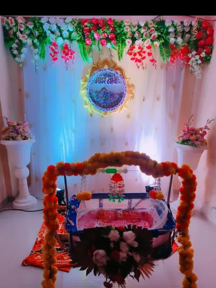A traditional home setup for a naming ceremony. The decor includes a decorated cradle, floral arrangements, and a backdrop with a sacred "Om" symbol.