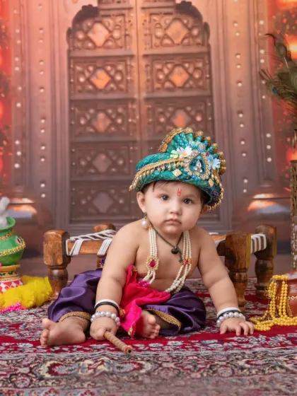 A direct and engaging portrait of a baby dressed as Krishna, holding his flute. The warm, glowing background with traditional Indian architecture creates a majestic setting.