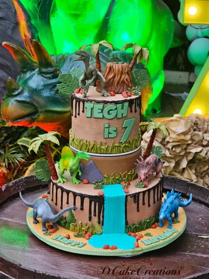 A roaring dinosaur-themed cake for Tegh's 7th birthday, set against a backdrop of jungle decor. The two-tier cake features a waterfall and various dinosaur species.