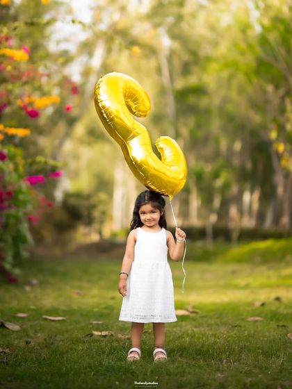 A happy girl holds her '3' balloon, celebrating her third birthday with an outdoor photoshoot.