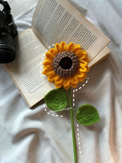 A single crochet sunflower styled with a book, showcasing its artistic and photogenic quality. It's a perfect prop for creating a cozy, aesthetic corner in your home.