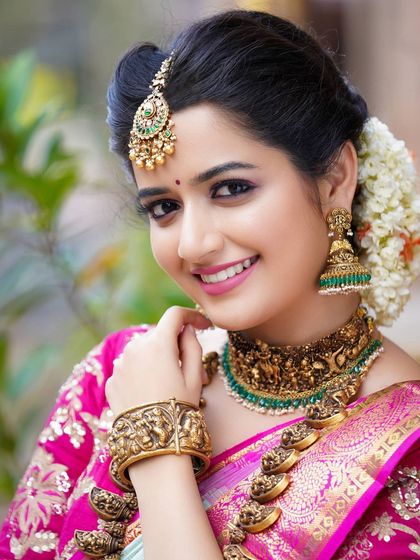 A full-view portrait of Ashika Ranganath, displaying the complete bridal look from the elegant saree drape to the detailed jewelry.