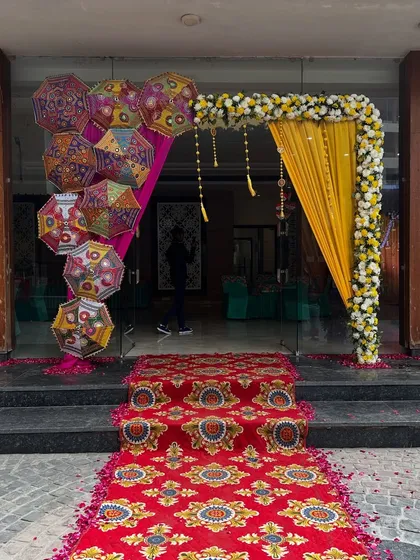 A beautiful entrance for a banquet hall, featuring a floral arch with yellow drapes, Rajasthani umbrellas, and a traditional patterned carpet.