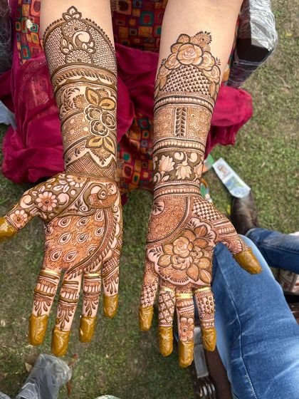 This outdoor shot shows how the rich color of the henna stands out. The design features a large peacock on one hand and a floral mandala on the other, both extending into full-arm patterns.
