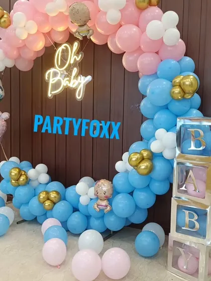 A gender reveal baby shower theme with a circular arch of pink and blue balloons, a neon "Oh Baby" sign, and transparent "BABY" blocks.