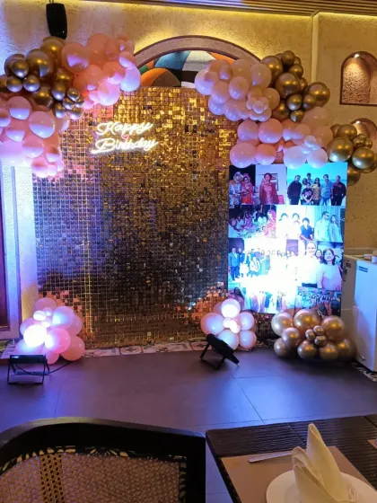 A personalized photo collage integrated into a birthday backdrop at a restaurant. The setup is framed by a gold sequin wall and a peach and gold balloon arrangement.