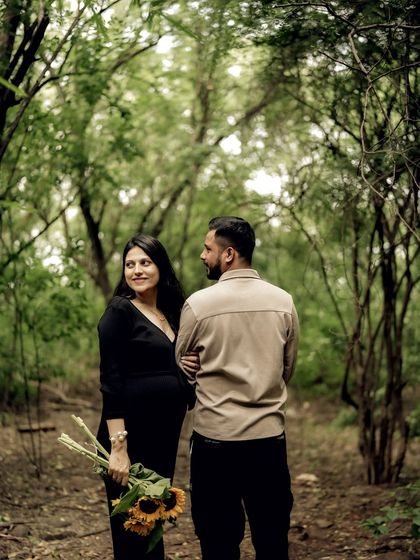 The couple stands together in a forest path, looking at each other with love. The natural frame of the trees adds to the intimacy of the scene.
