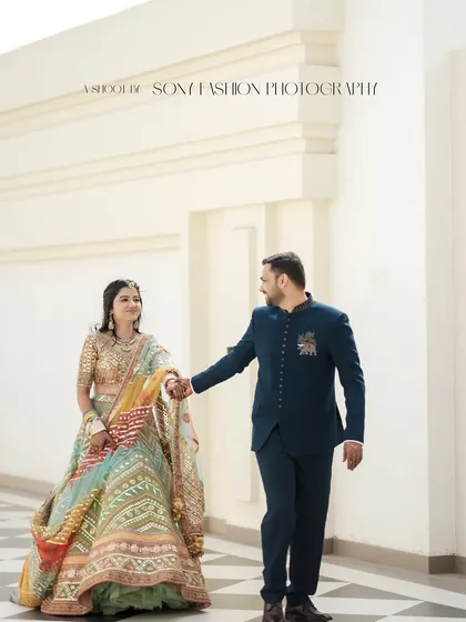 The couple walks hand-in-hand through a pristine white corridor with a checkered floor. This shot has a clean, elegant feel, highlighting the bride's stunning multi-colored lehenga.