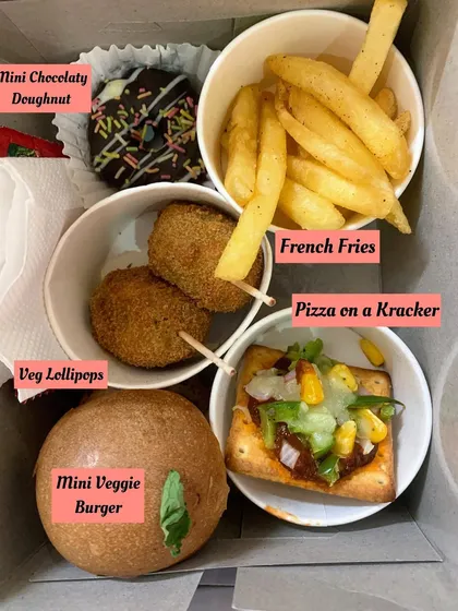 A close-up of a perfectly portioned snack box for a kid's party. It includes a mini veggie burger, veg lollipops, french fries, pizza on a cracker, and a mini chocolate doughnut, a complete and happy meal for any child.