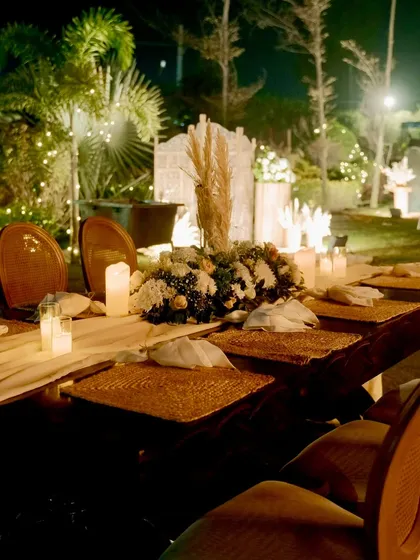 An intimate and rustic dining table setup for an outdoor celebration. We used a wooden table, woven placemats, and a centerpiece of white flowers and pampas grass.