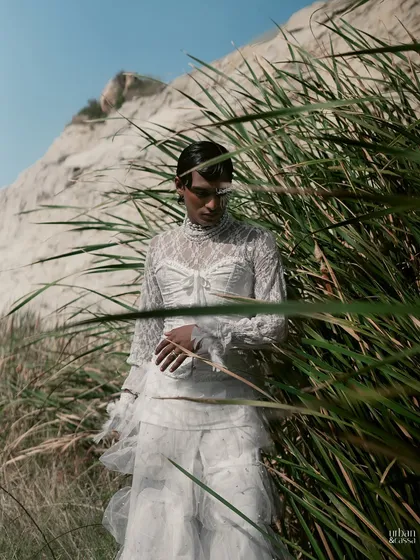 The model is framed by tall grass, adding a natural element that contrasts with the highly stylized makeup and hair.