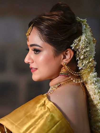 A stunning profile shot that highlights the intricate hairstyle adorned with fresh jasmine flowers. The makeup is kept classic and clean to balance the elaborate hair and jewellery.