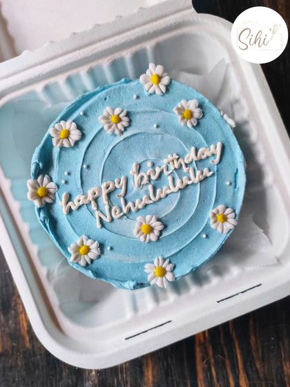 A beautiful blue bento cake with a spiral frosting pattern. The design is finished with a circle of fondant daisies and a "Happy Birthday" message in lovely script.