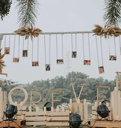 A creative photo backdrop for an anniversary, featuring a 'FOREVER' sign and hanging photos of the couple's journey, framed by rustic wooden elements and pampas grass.