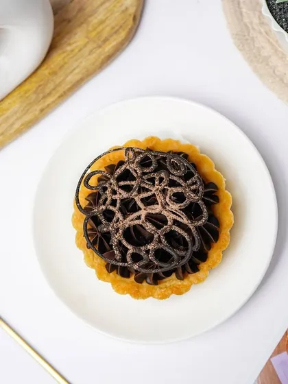 An elegant chocolate tart with intricate piping on top, shot from above on a white plate. This minimalist, bright setup is perfect for showcasing artisanal pastries and desserts.
