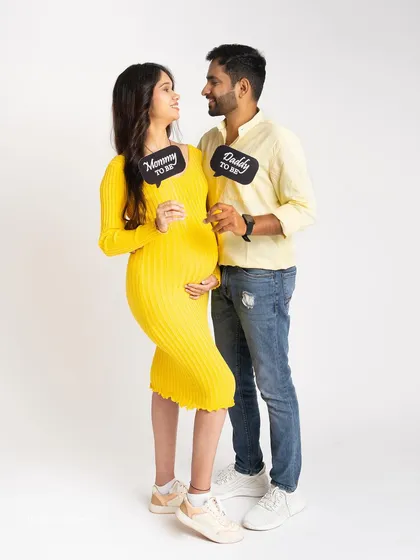A fun and casual shot of an excited couple. They hold up "Mommy to be" and "Daddy to be" signs, dressed in bright, coordinated yellow outfits against a clean white background.