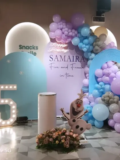 A 'Frozen' themed birthday party for Samaira's 5th birthday. The setup includes life-sized cutouts of Elsa and Olaf, a marquee number '5', and a balloon cascade in icy blue, purple, and silver.