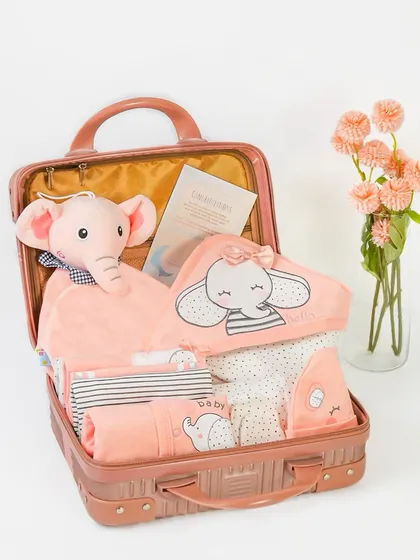 This shot reveals the contents of a baby girl hamper packed in a vintage-style suitcase. It showcases a complete gift set, including clothes, a soft toy, and a blanket.
