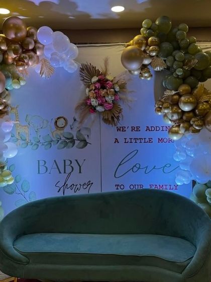 An elegant baby shower setup with an olive green and gold balloon arrangement. The backdrop features the message 'We're adding a little more love to our family', creating a perfect photo spot.