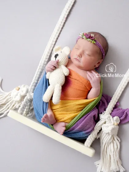 A rainbow baby. This beautiful portrait features a baby wrapped in rainbow colors, holding a teddy bear while resting on a swing.