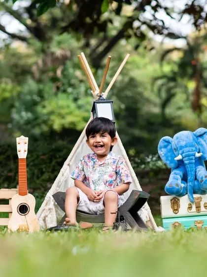A smiling boy sits in front of a teepee during his outdoor third birthday photoshoot, surrounded by fun props.