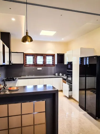This open-concept modular kitchen is designed for both style and efficiency. It features a sleek two-tone design with high-gloss white and dark wood finish cabinets, a functional island counter, and integrated appliances, all illuminated by a combination of recessed lights and a statement pendant lamp.