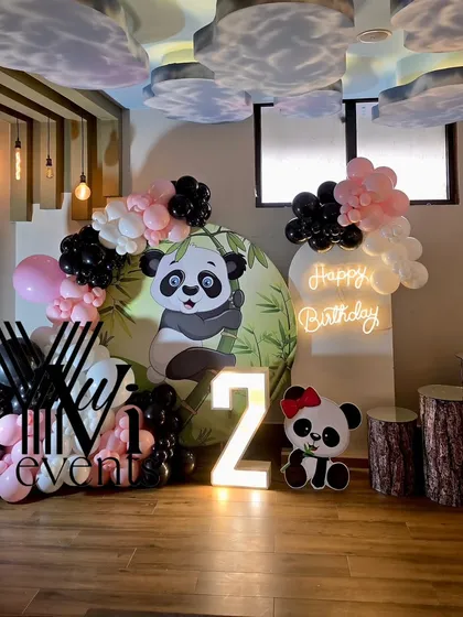 A cute panda-themed second birthday party setup, with a custom backdrop, black, white, and pink balloons, and a light-up number '2'.