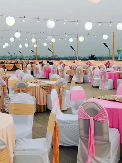A beautiful rooftop seating setup for a reception. The pink and peach chair covers, combined with the overhead paper lanterns, create a festive and elegant ambiance against the city skyline.
