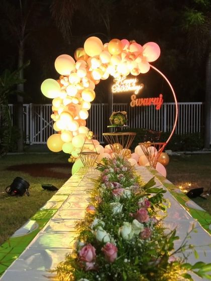 A breathtaking outdoor birthday setup at night, with a long, flower-lined walkway leading to a beautifully lit balloon arch and neon sign.