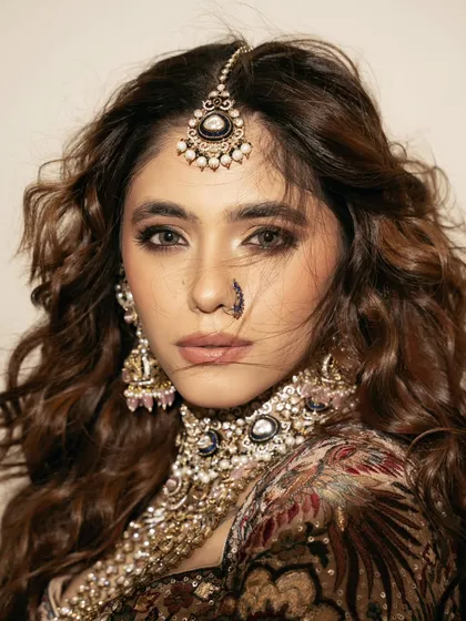 A tight close-up on the bride's face, focusing on the intricate details of the maang tikka, nose ring, and choker. The wind-blown hair adds a touch of softness and movement.