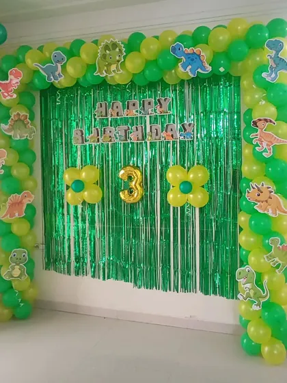 A dinosaur-themed party setup for a third birthday. I used a green and yellow balloon arch decorated with various dinosaur cutouts, set against a green shimmer curtain to create a prehistoric jungle vibe.
