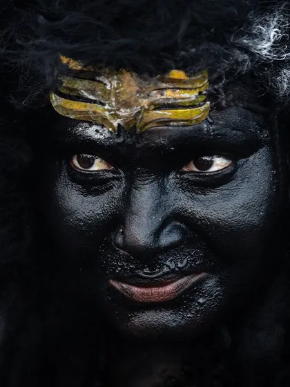 An intense close-up of a devotee's face, painted black with a tripundra on the forehead, capturing the fierce and focused energy of the divine character they embody.