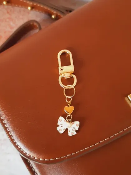 A simple and chic bag charm featuring a white enamel bow with polka dots and a tiny heart. It's perfect for adding a subtle, stylish touch to a classic brown leather bag.