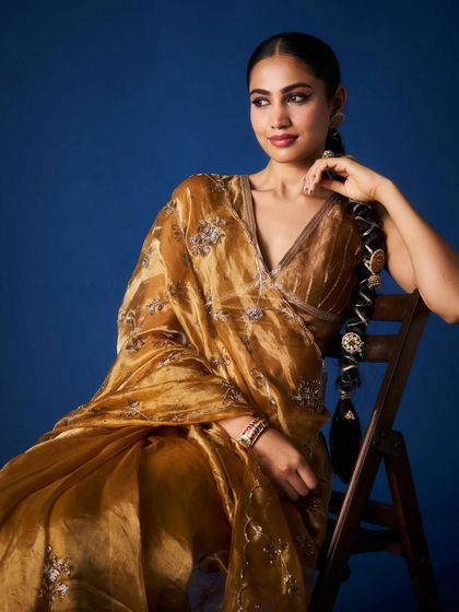 A model sits gracefully in a molten gold tissue saree with silver floral embroidery, her look completed with a traditional braided hairstyle.