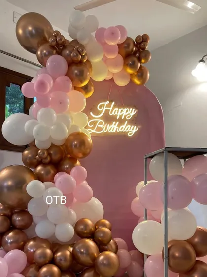 A chic 'Happy Birthday' decoration in pink, white, and bronze. The arched backdrop and balloon garland can be customized in any color to match your party's theme.
