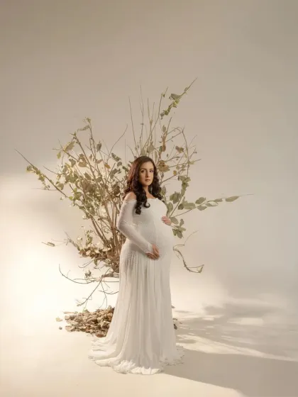 A fine art portrait in our studio. The mother-to-be, in a flowing white gown, stands before a rustic, artistic installation of branches, creating a beautiful contrast between soft elegance and natural texture.