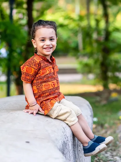 A sweet portrait of a two-year-old boy in a traditional print kurta, smiling while sitting on a rock outdoors.