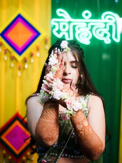 A beautiful photo of a bride with her dark henna stain, adorned with floral jewelry. The "Mehndi" neon sign in the back sets the perfect mood.