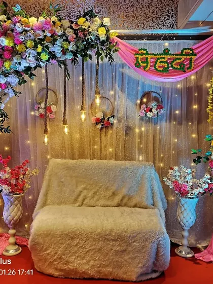 A festive Mehndi stage setup combining a lush floral arch with bright pink drapes and fairy lights. The 'Mehndi' sign in Hindi adds a traditional touch to this vibrant and colorful decoration.