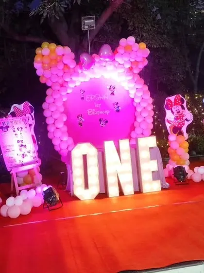A Minnie Mouse themed birthday party shown at night. The setup, featuring a pink balloon backdrop and light-up 'ONE' letters, is illuminated with spotlights to create a magical effect for an evening celebration.