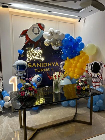 A space-themed setup for Sanidhya's first birthday, featuring a rocket ship, astronaut cutouts, and a "Happy Birthday" neon sign.