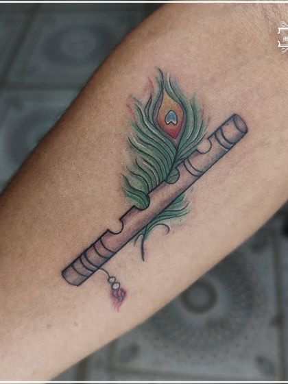 A colored tattoo of a peacock feather and a flute (bansuri), symbols of Lord Krishna. The vibrant colors make this spiritual and nature-themed piece truly stand out.