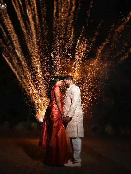 A couple shares an intimate moment, their faces illuminated by a brilliant explosion of fireworks behind them. This night shot from a wedding celebration captures the magic and energy of the festivities with dramatic lighting.